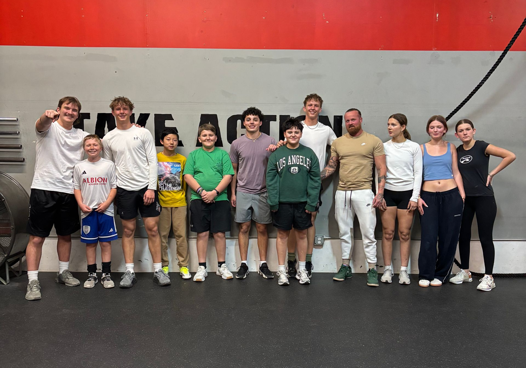 A group of seven people posing in a gym, with a shirtless man centered in the middle, under overhead warehouse lighting.