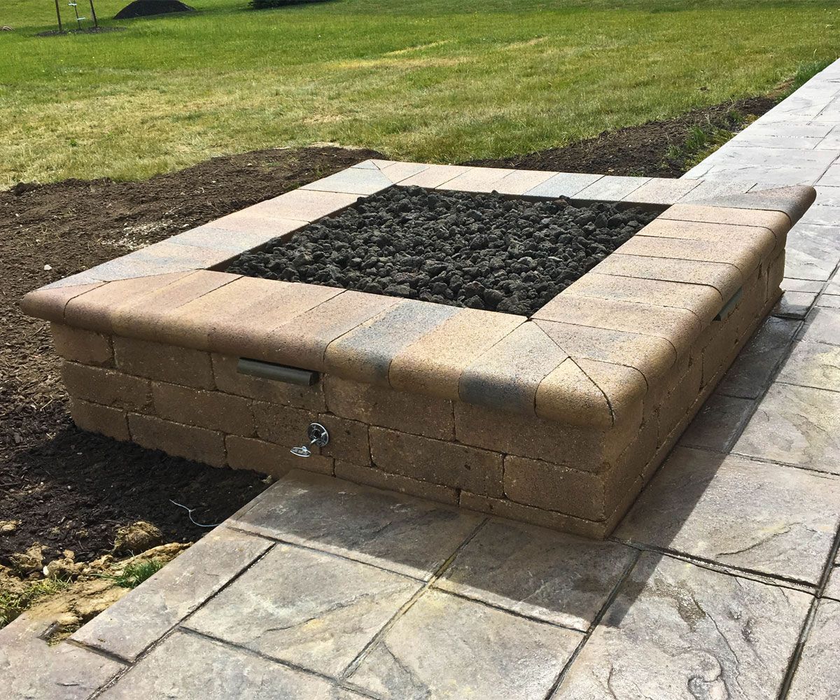 Square Unilock wall block gas fire pit with coping, outdoor landscape lighting, and black fire lava rock in Cuyahoga County backyard.
