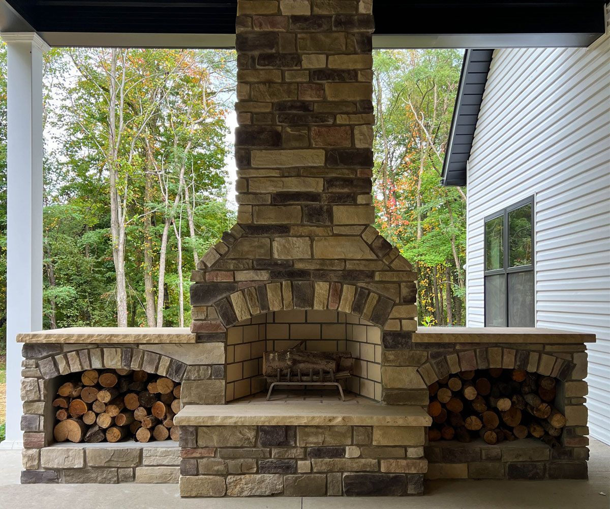 Stone-clad outdoor fireplace with extended chimney and fire wood boxes on a covered Medina paver patio