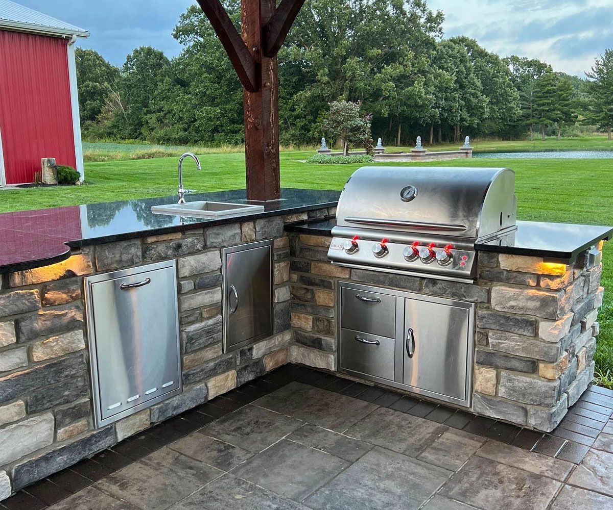 Multi-tiered L-shape outdoor kitchen covered under pavilion in Northeast Ohio backyard on paver patio