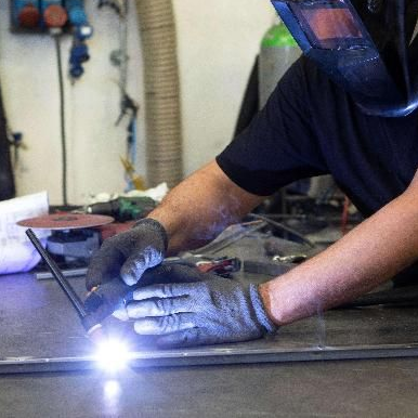 Saldatore con casco e guanti protettivi che salda metallo su un banco da lavoro, arco luminoso in officina