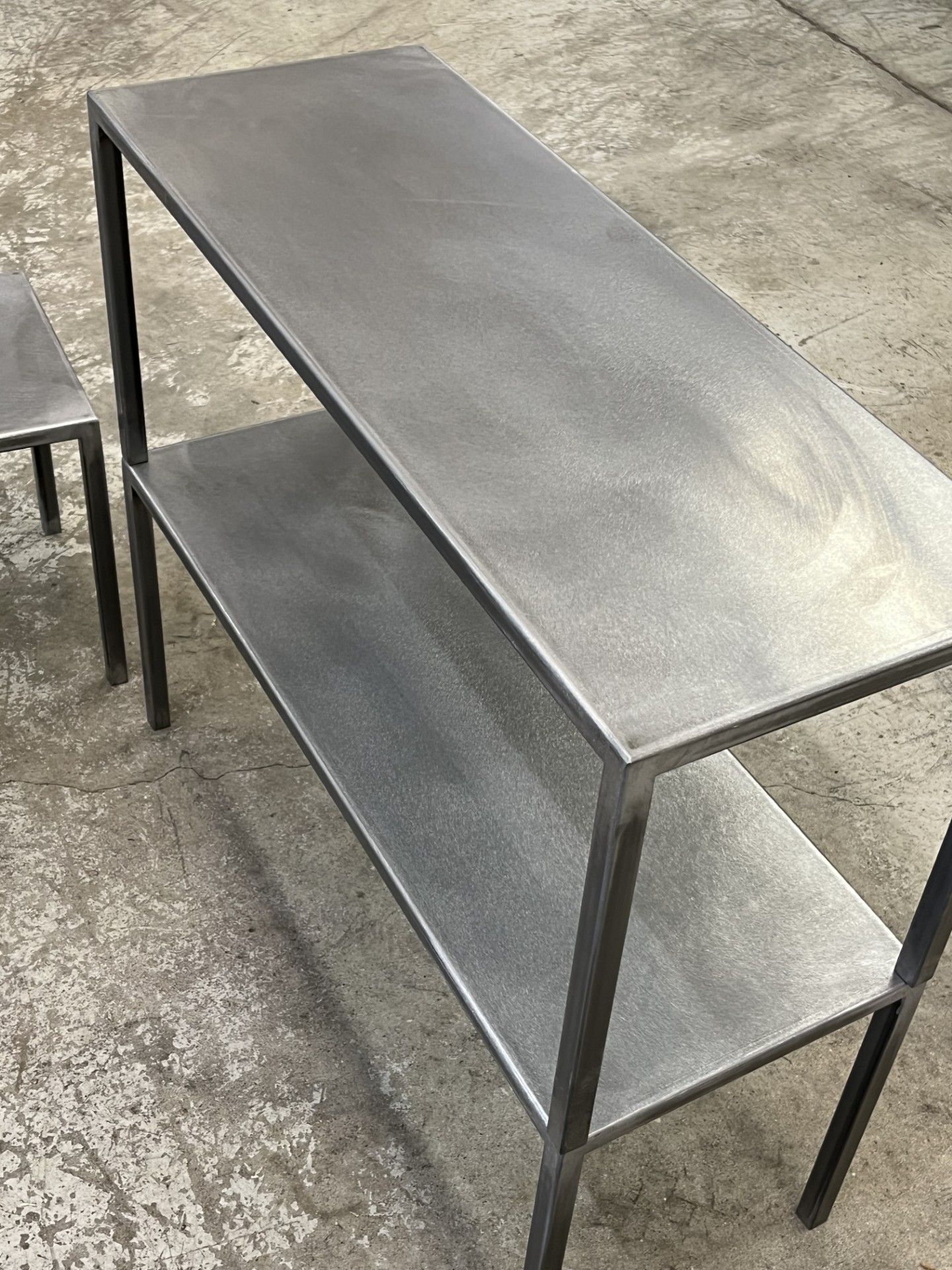 Carrello portautensili a due ripiani in metallo con struttura scura e ripiani grigi su pavimento in cemento.