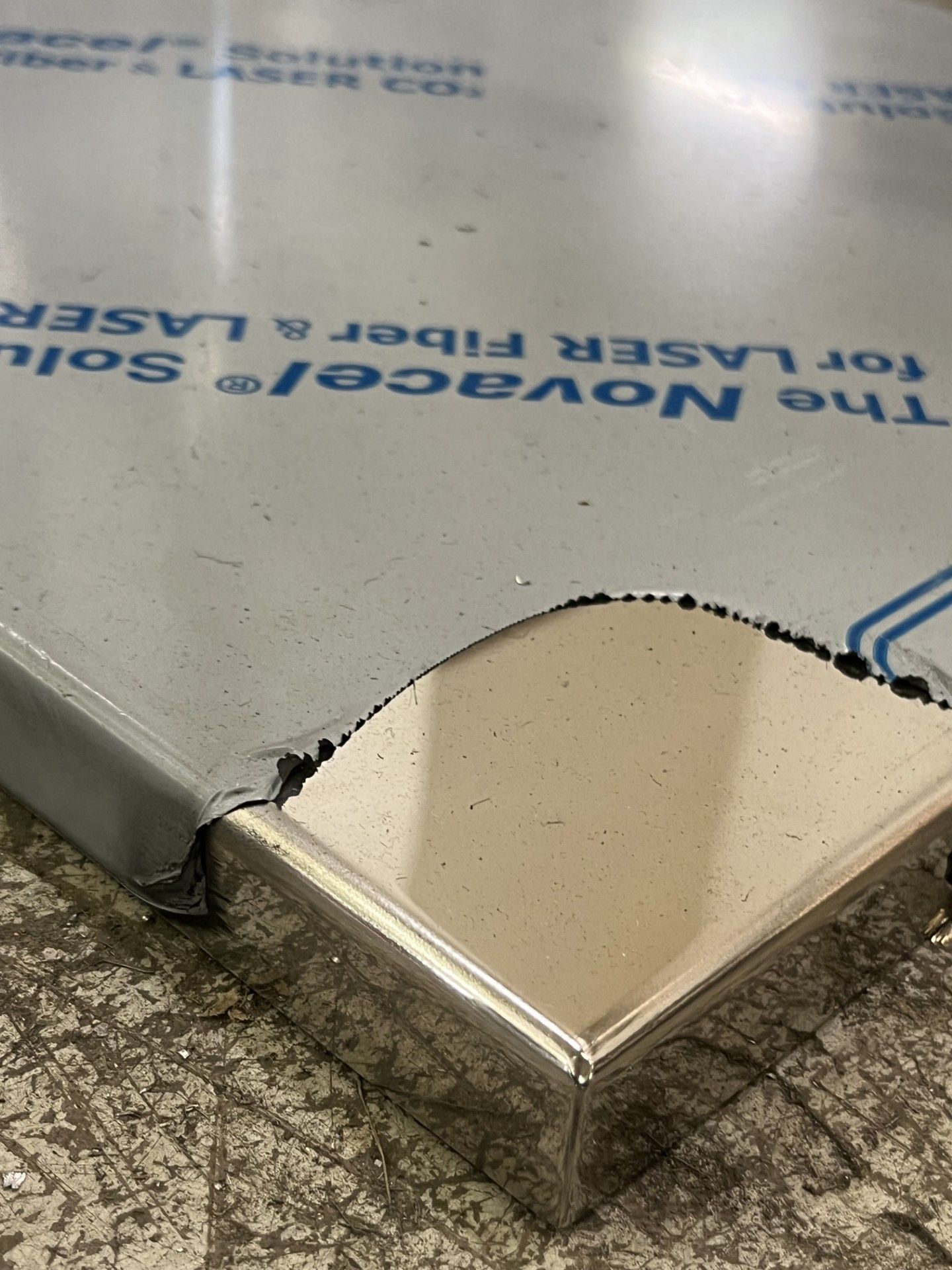 Primo piano di un angolo di pavimento metallico danneggiato con un bordo scheggiato e un foglio di plastica protettivo nelle vicinanze.