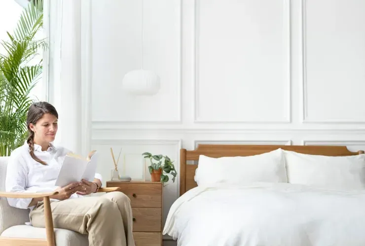 Woman reading in a bright bedroom beside a neatly made white bed