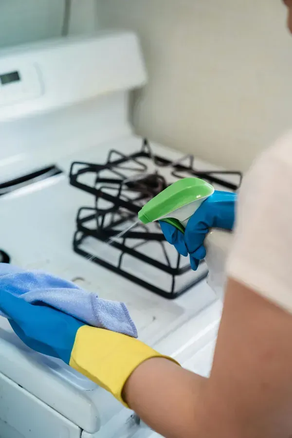 Gloved hands cleaning a white stove with a green sponge and spray bottle.