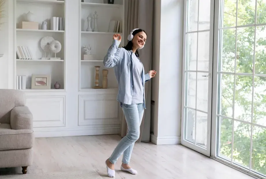 Woman in a light blue shirt dancing in a bright living room by large windows.