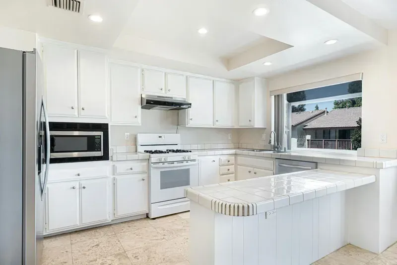 Bright white modern kitchen with island, white cabinets, stove, and large window.