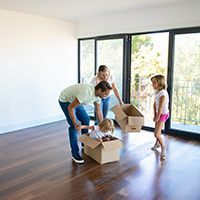 Family unpacking boxes in a bright empty room with large windows and wood floors
