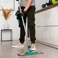 Person mopping a kitchen floor with a turquoise spray mop, wearing black pants and white sneakers