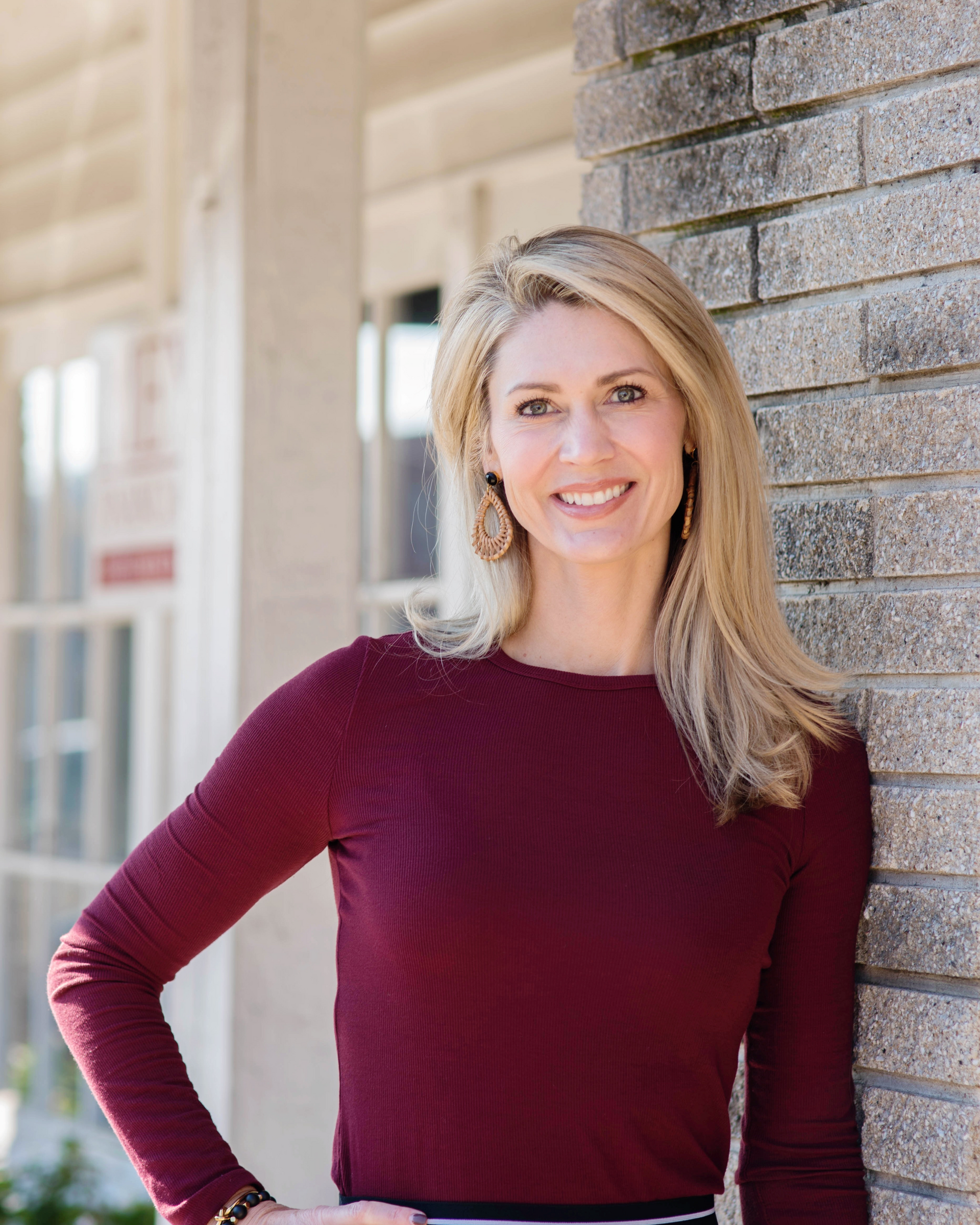 Smiling woman in a burgundy top leaning against a brick wall outdoors
