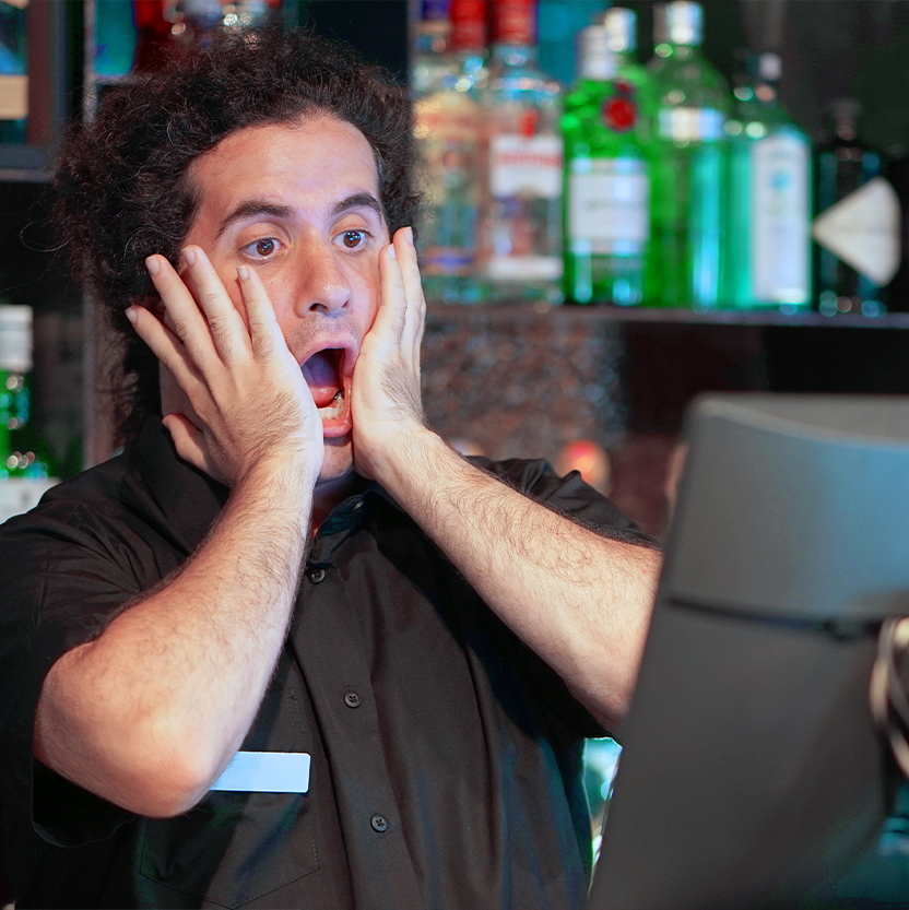 Shocked bartender with hands on cheeks behind a bar, looking at a monitor.