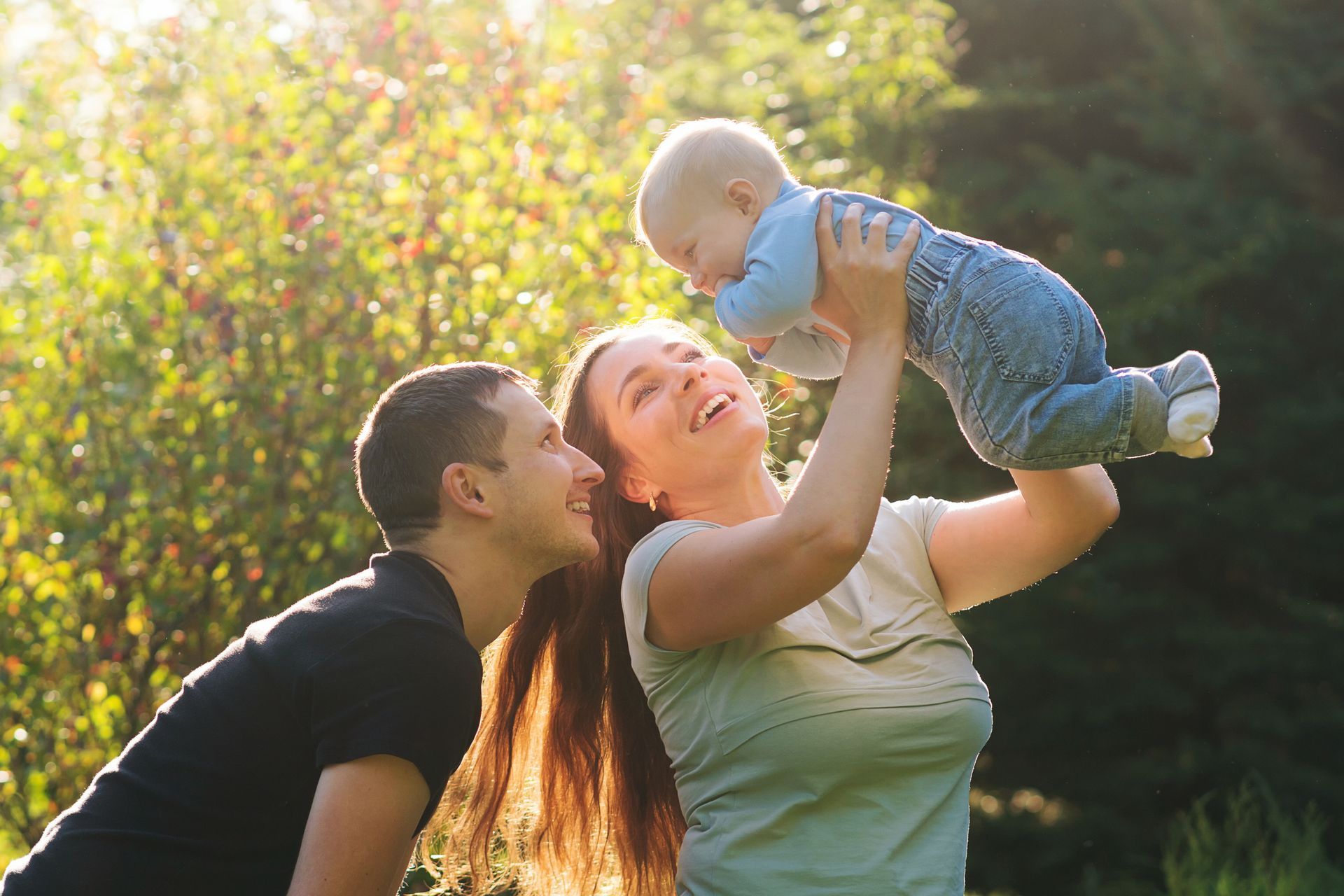 Two adults lifting a baby outdoors in a sunlit park