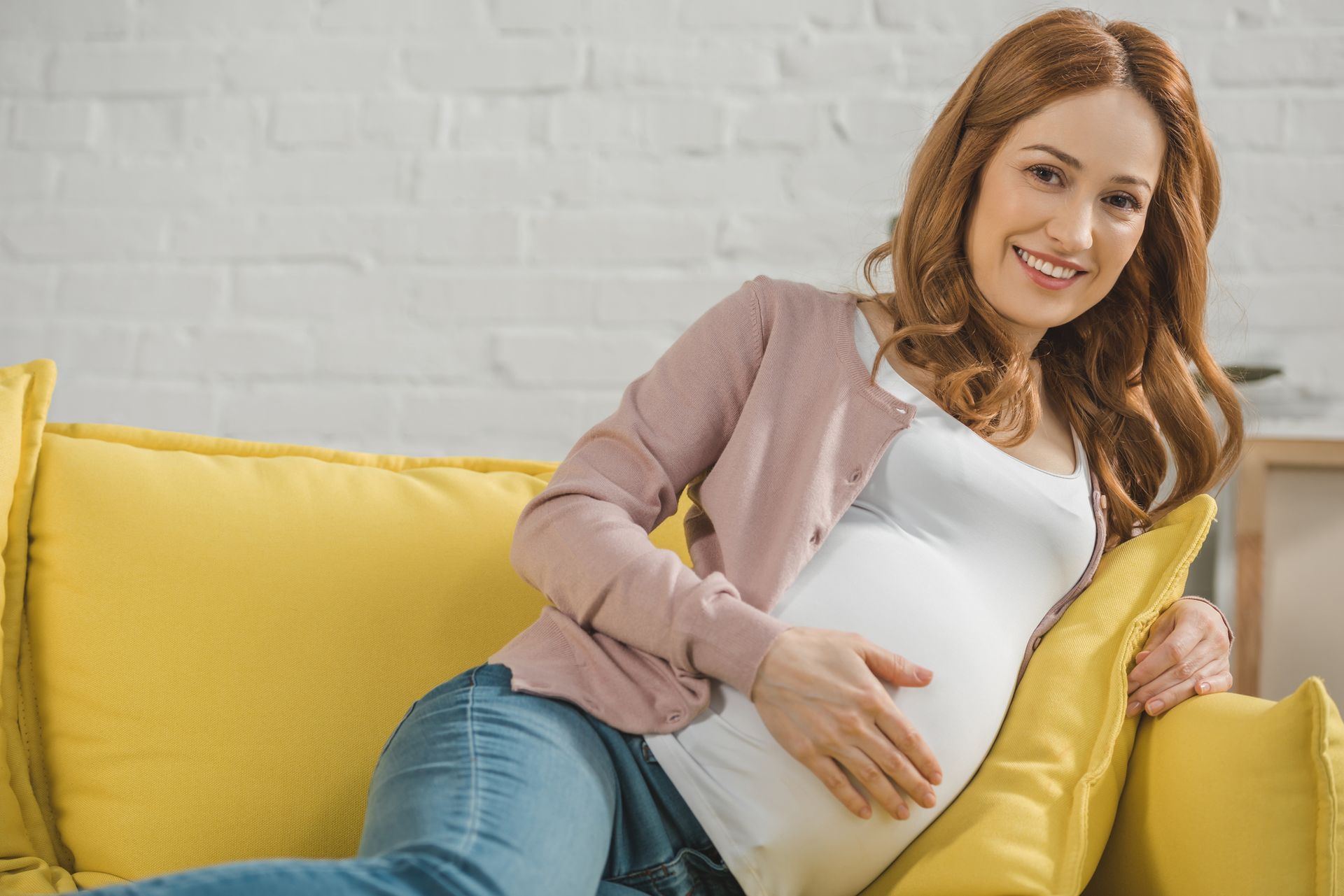 Pregnant woman reclining on a yellow couch, smiling and resting her hand on her belly