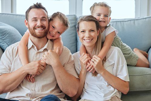 Family of four smiling on a couch in a bright living room, with the parents holding their children close.