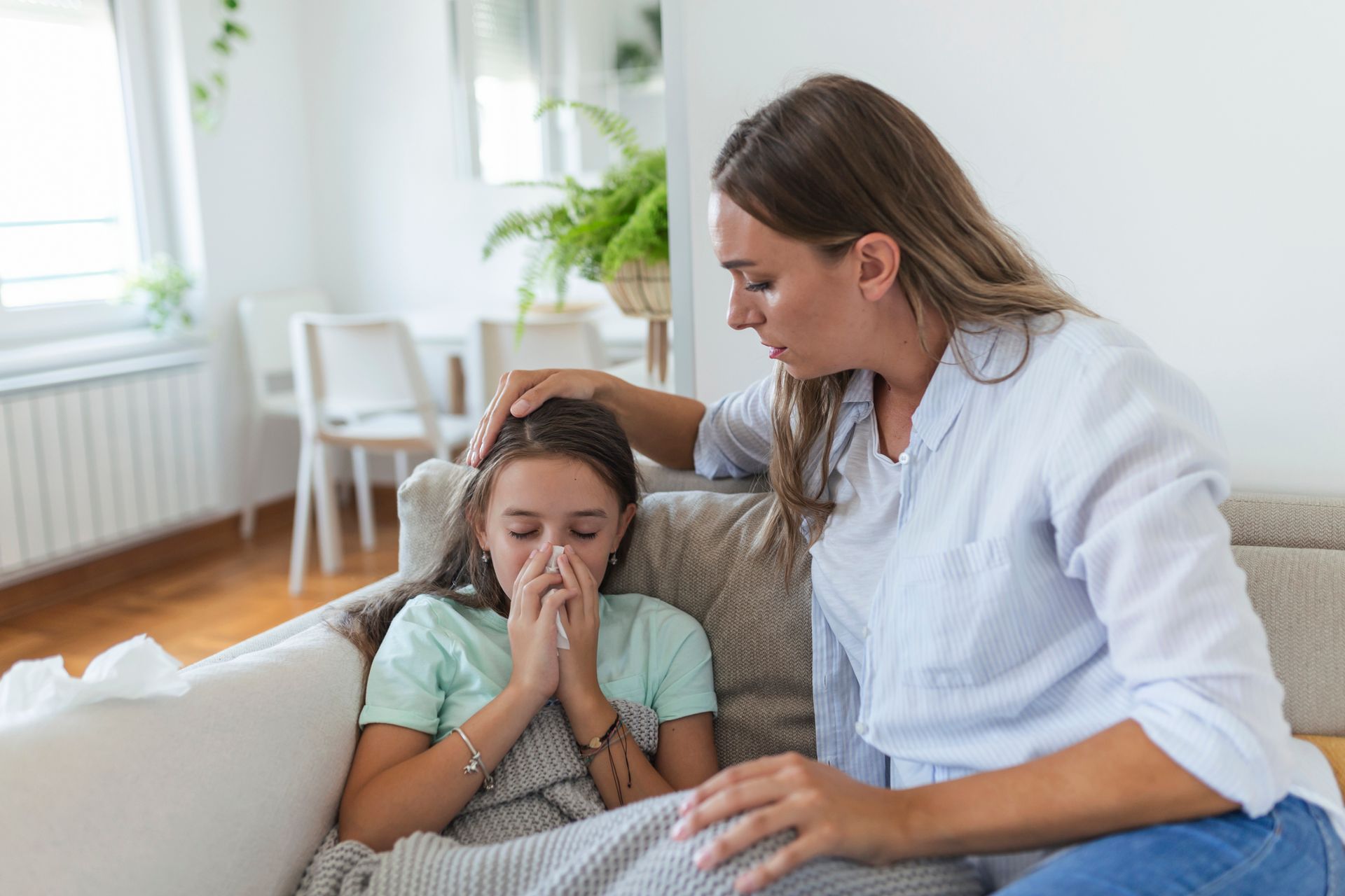 Woman comforting a sick child on a sofa in a bright living room, child holding tissues and looking distressed