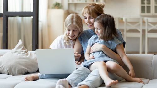 Three people sit on a couch, smiling and looking at a laptop in a bright living room.