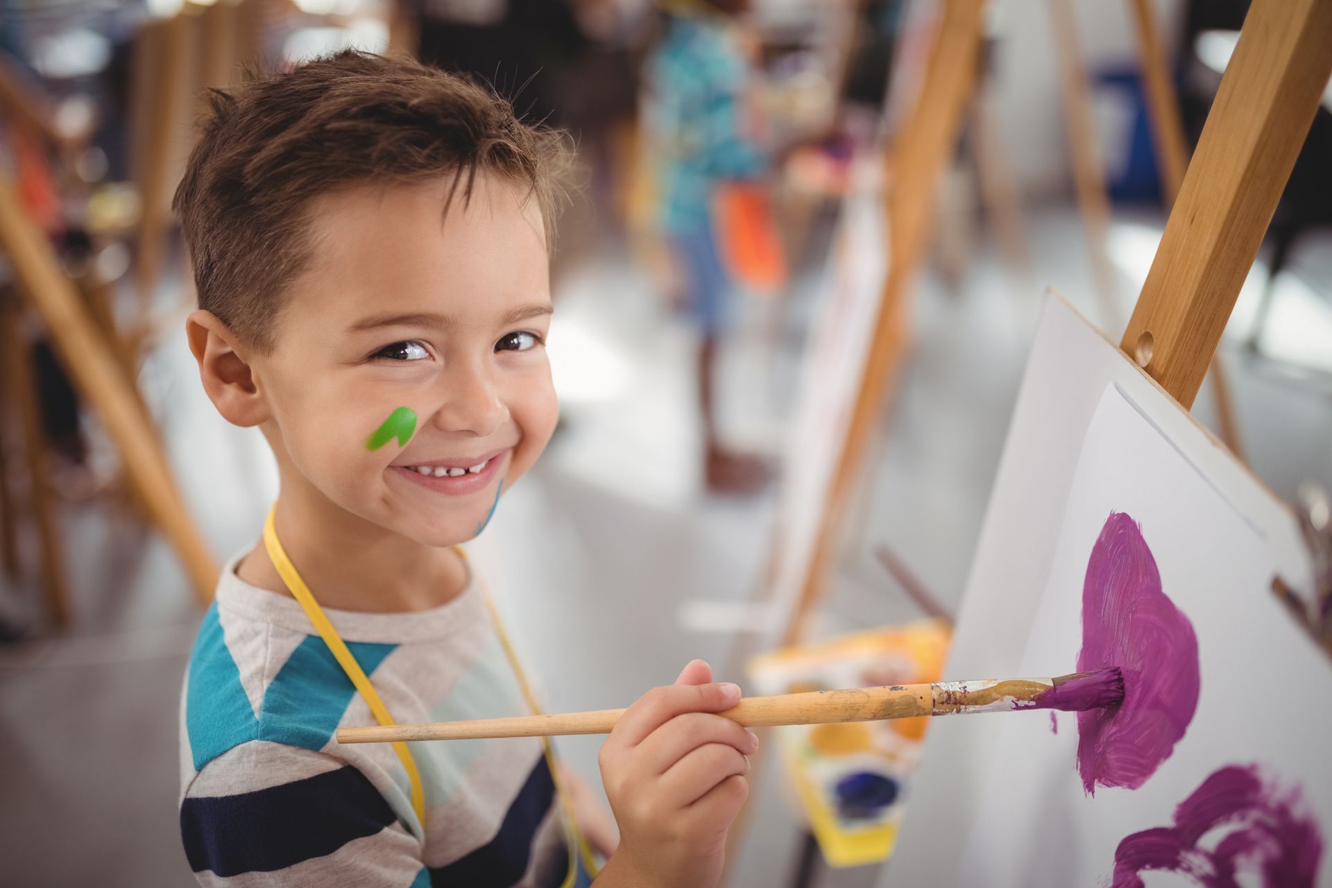 Smiling child painting on a canvas with a brush, colorful paint on cheek, in an art studio