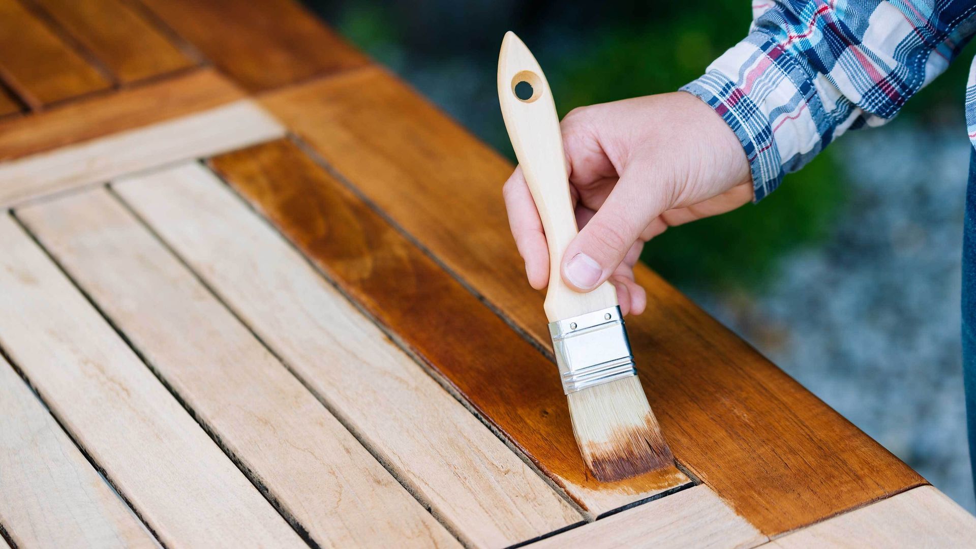 Hand painting a wooden outdoor table with a brush, applying stain to a slatted surface.