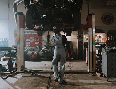Mechanic stands under a raised car in a dim auto repair shop, facing the vehicle.