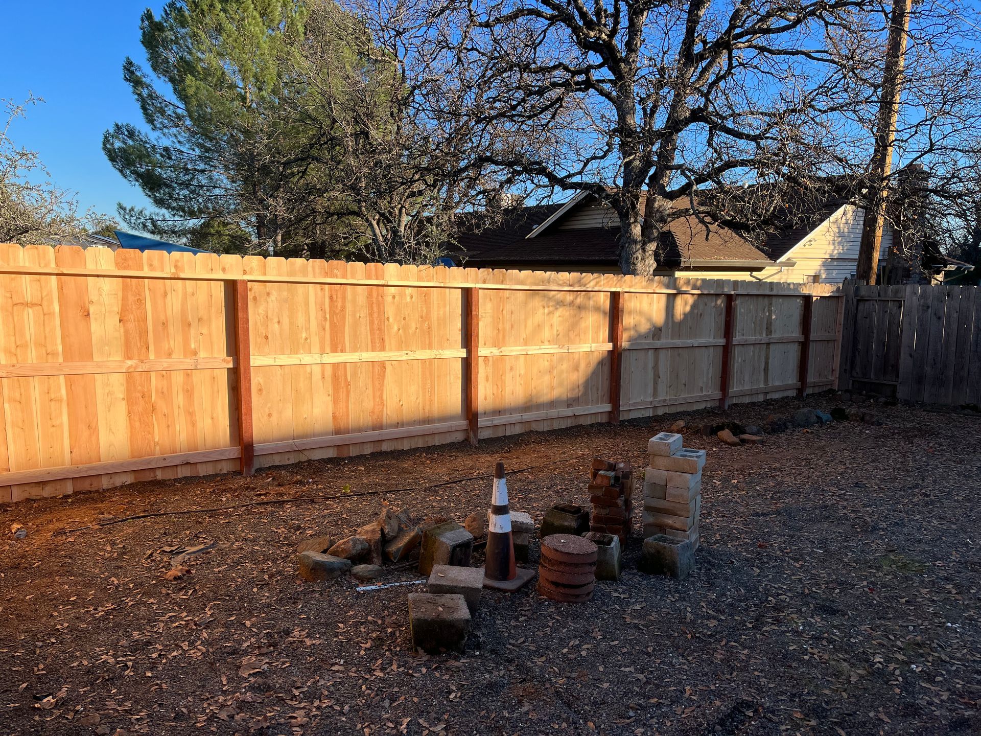 Wooden backyard fence with stacked bricks and a small fire pit on gravel under bare trees.
