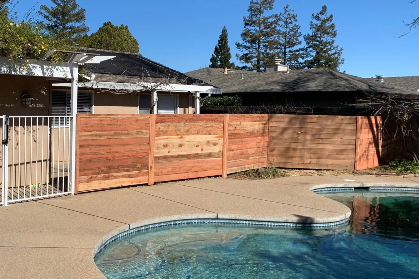 Backyard pool with a wooden fence and single-story house under a clear blue sky
