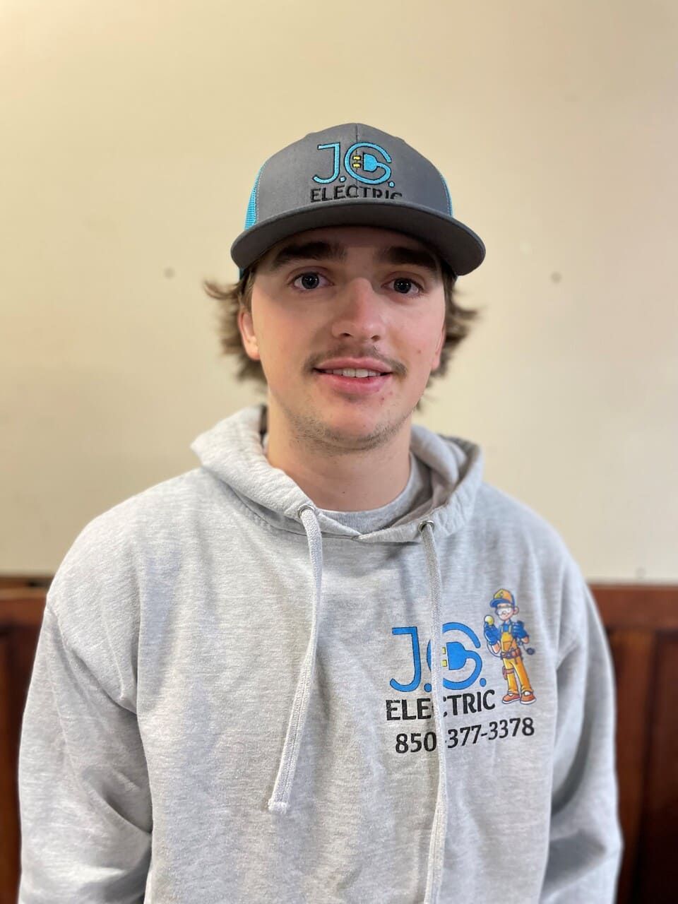 A person wearing a grey JC Electric hoodie and matching baseball cap poses against a plain beige wall.