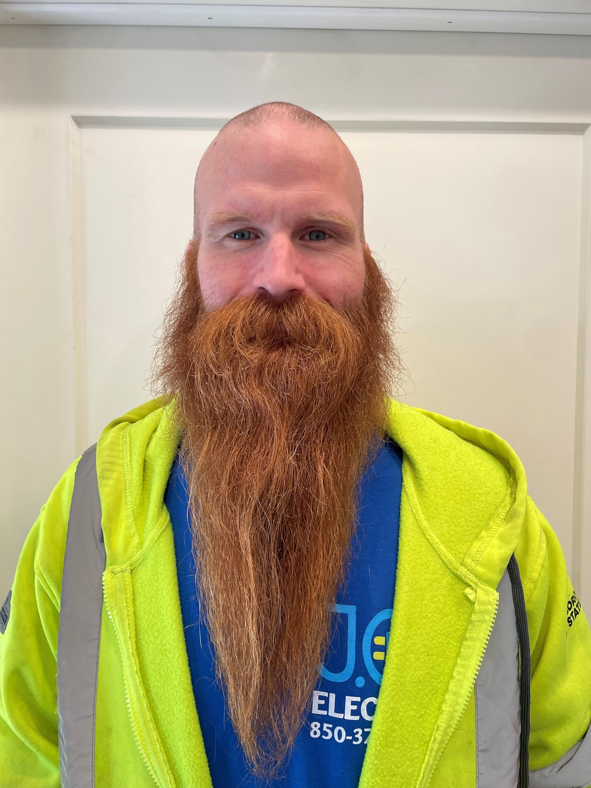 A person with a long, thick, reddish-brown beard, wearing a bright yellow high-visibility jacket over a blue shirt.