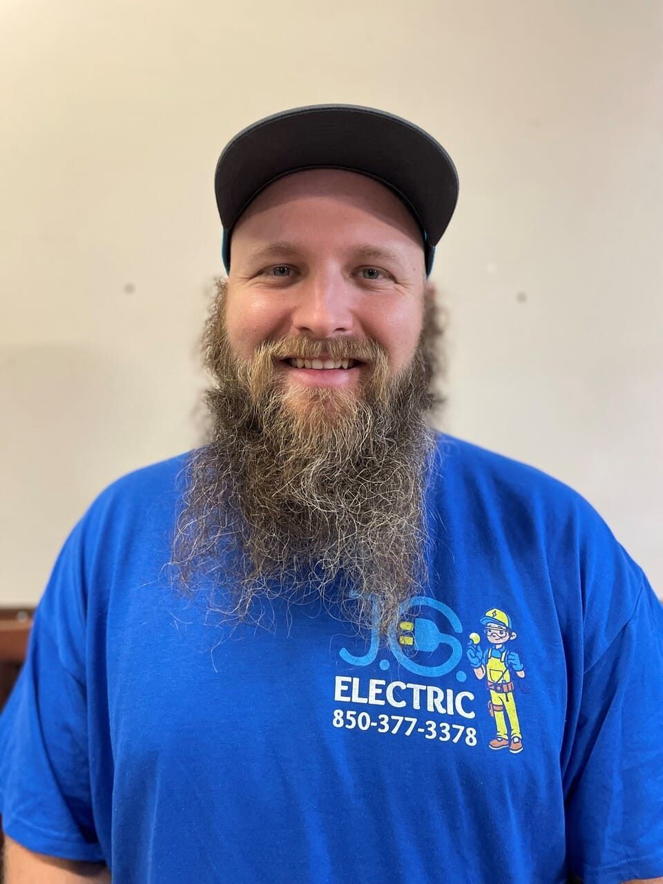 A person with a long beard and a black cap smiles while wearing a blue J.G. Electric T-shirt against a plain background.