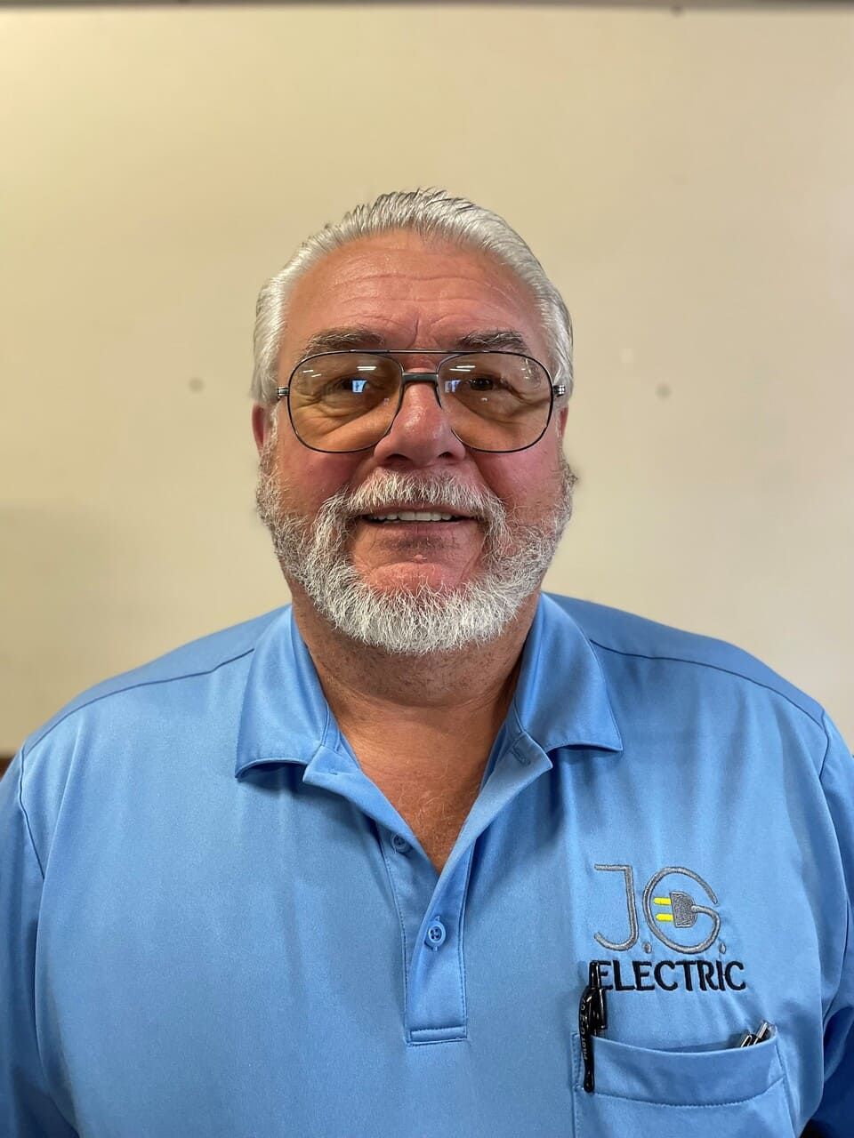 A person with a grey beard and glasses wears a light blue polo shirt with a