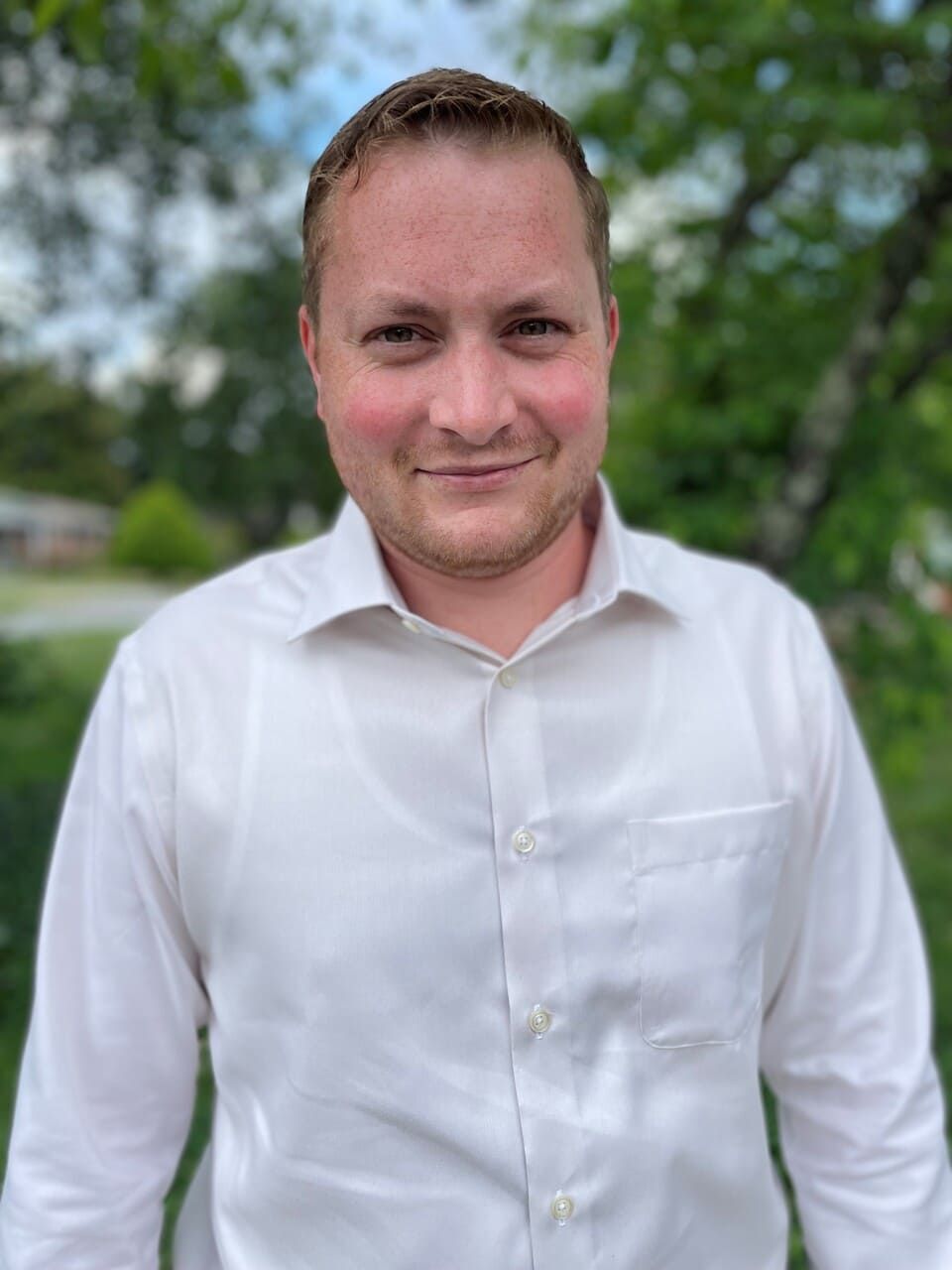 A person in a light-colored, long-sleeved button-down shirt smiling slightly, outdoors with trees in the background.