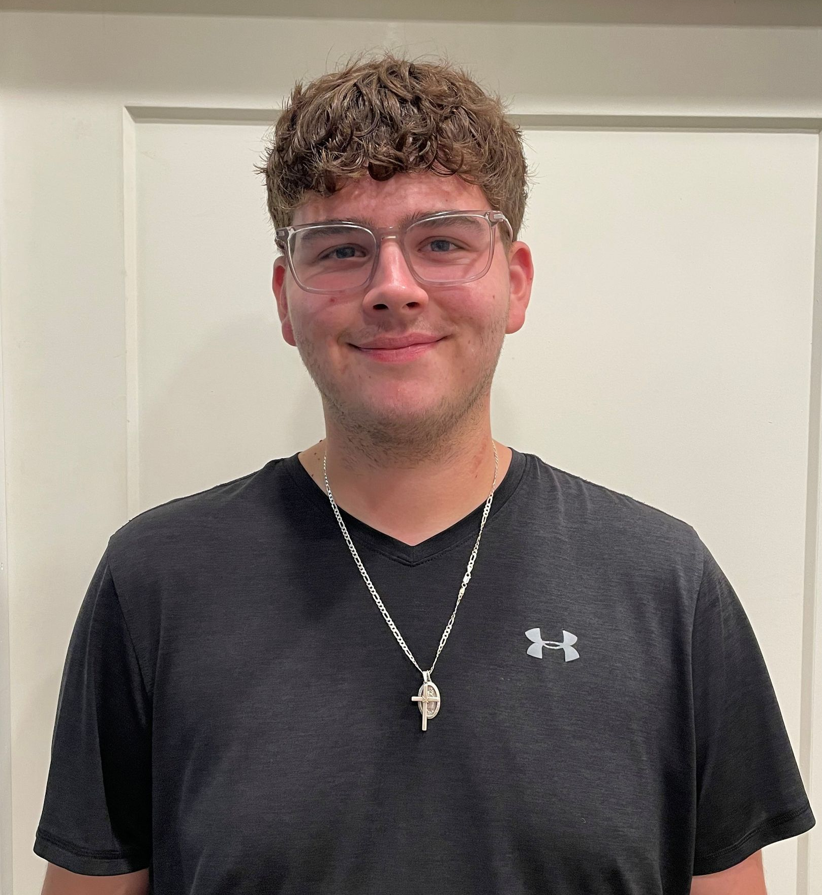 A person with curly hair and glasses, wearing a dark gray v-neck shirt and a silver pendant necklace, smiling at the camera.