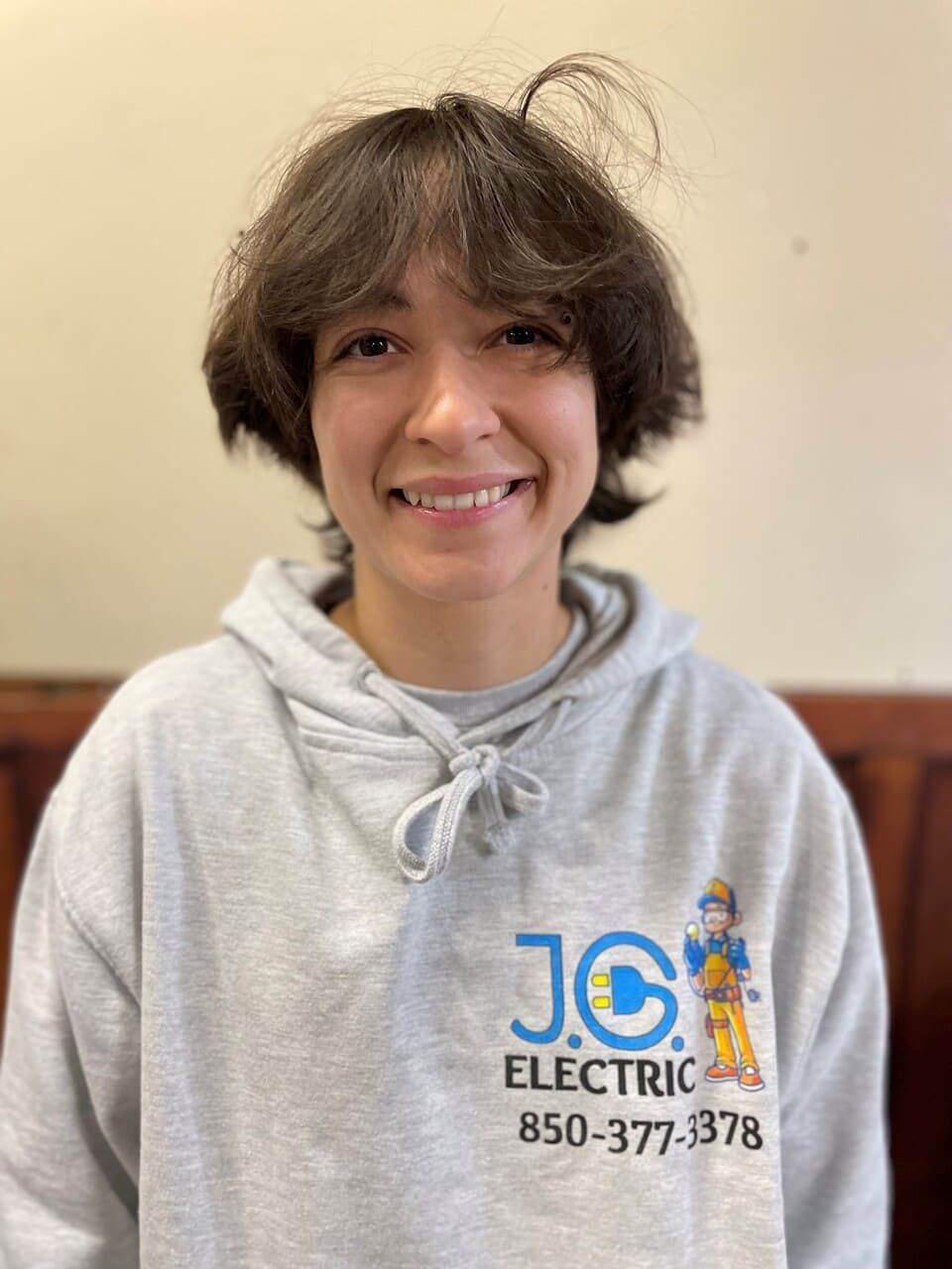 A person with short, brown hair smiles at the camera, wearing a gray hooded sweatshirt with a business logo on the chest.