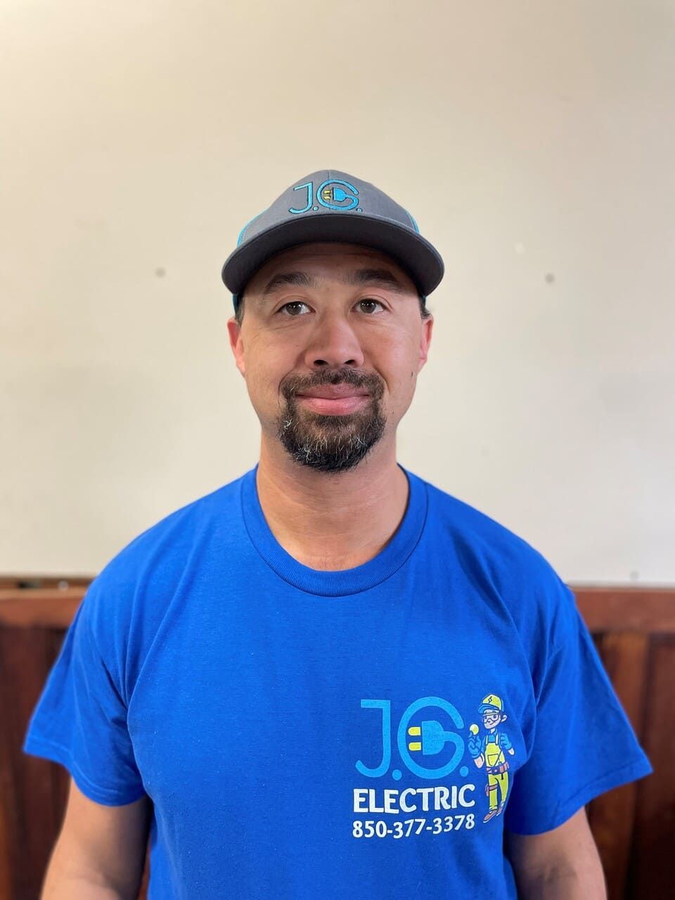 A person wearing a blue J.G. Electric t-shirt and grey cap stands against a plain background.