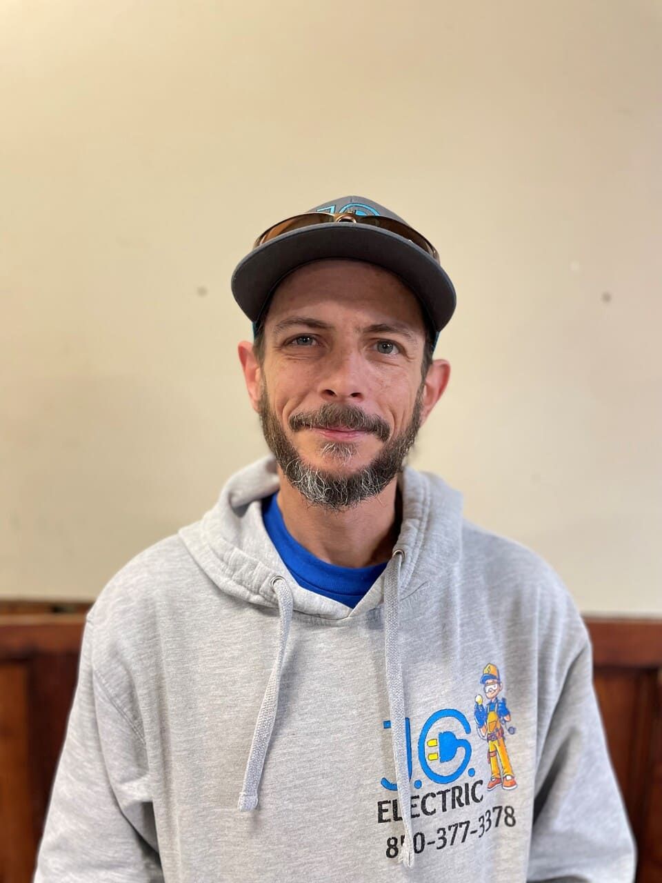 A man with a beard, wearing a baseball cap and a gray J.C. Electric hooded sweatshirt, against a plain background.