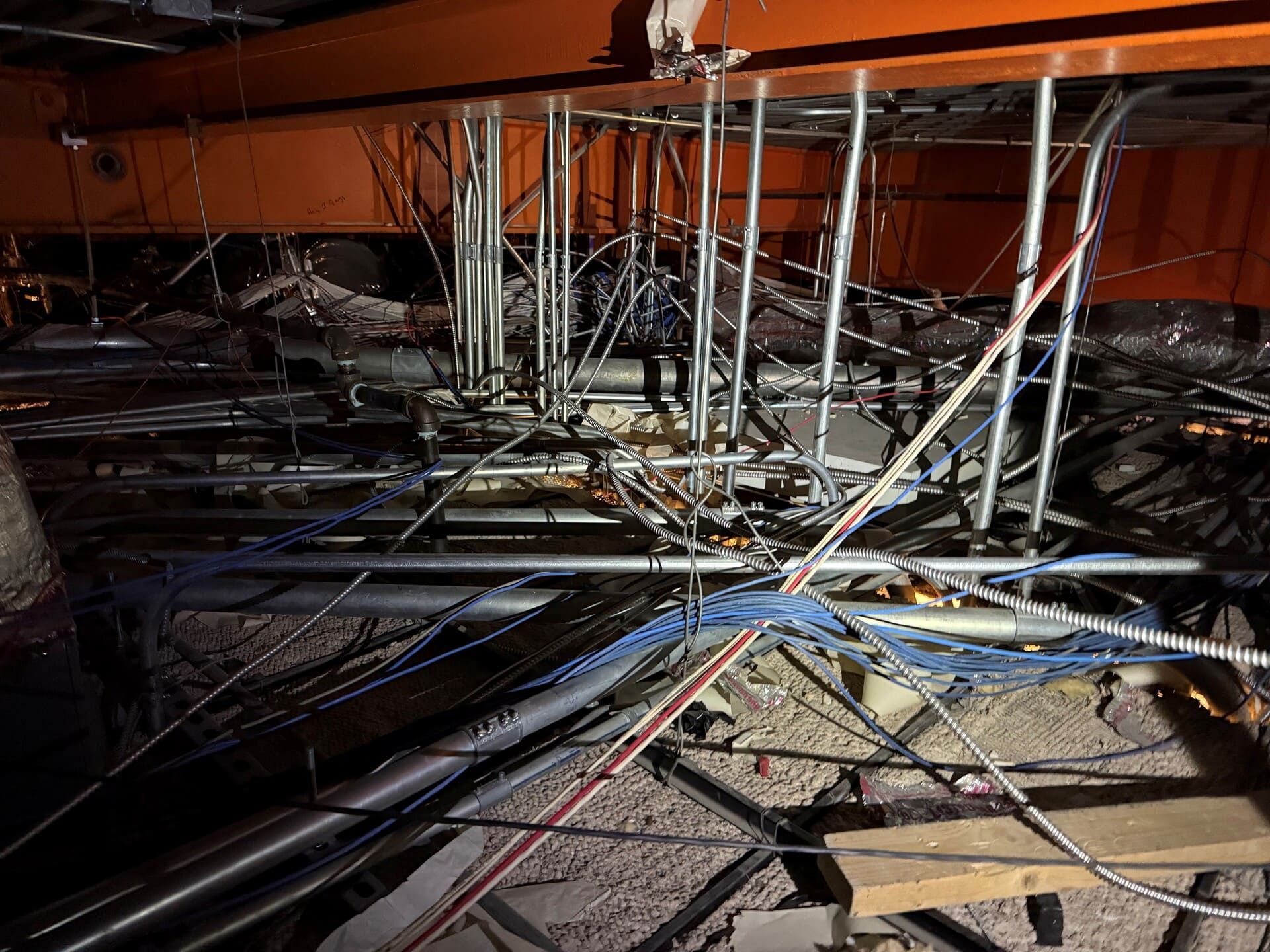 A ceiling plenum space showing a dense web of wires, metal conduits, and pipes mounted below a large orange structural beam.