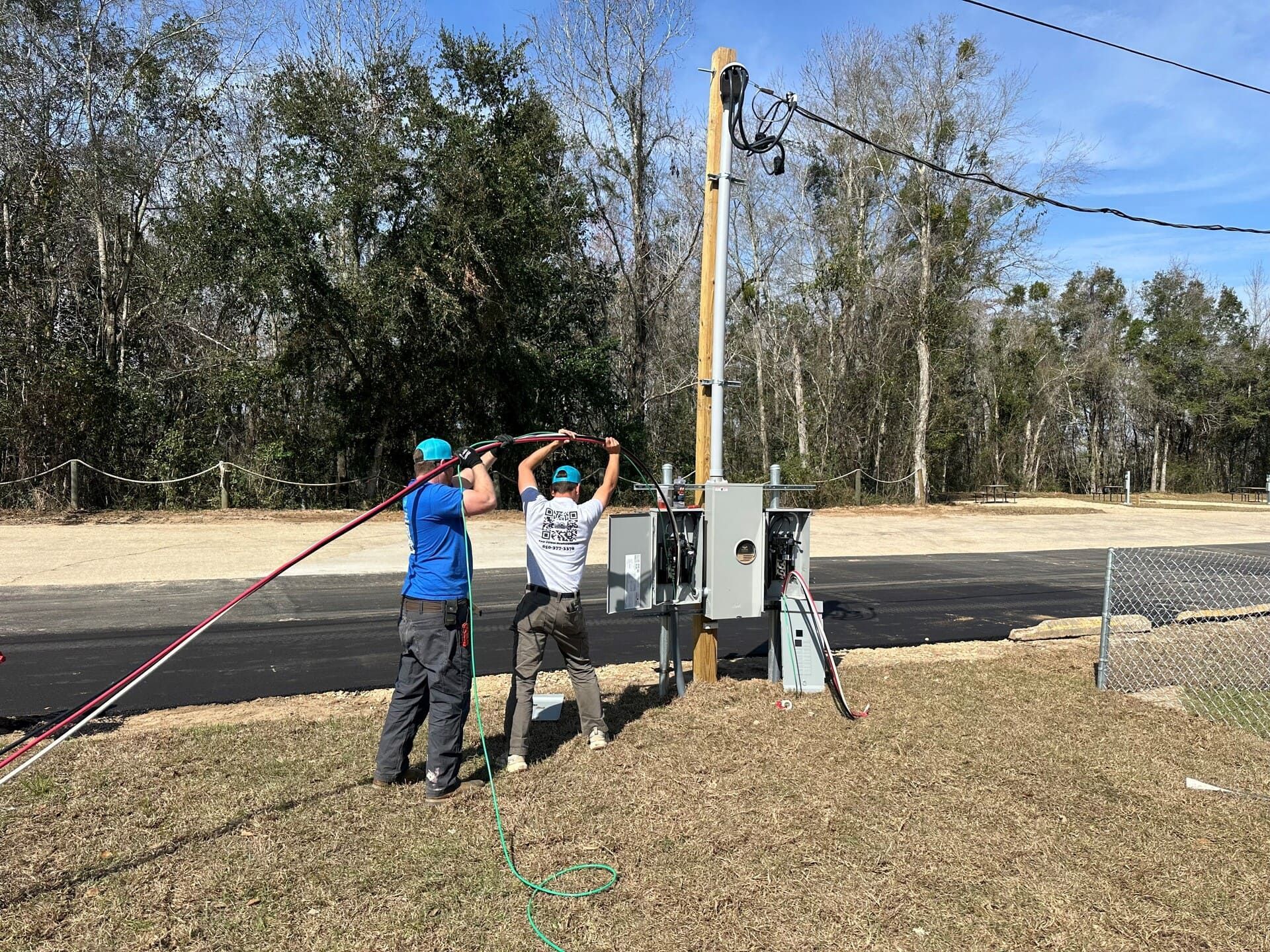 Two workers in blue and white shirts install a power cable into an electrical box near a utility pole and paved road.