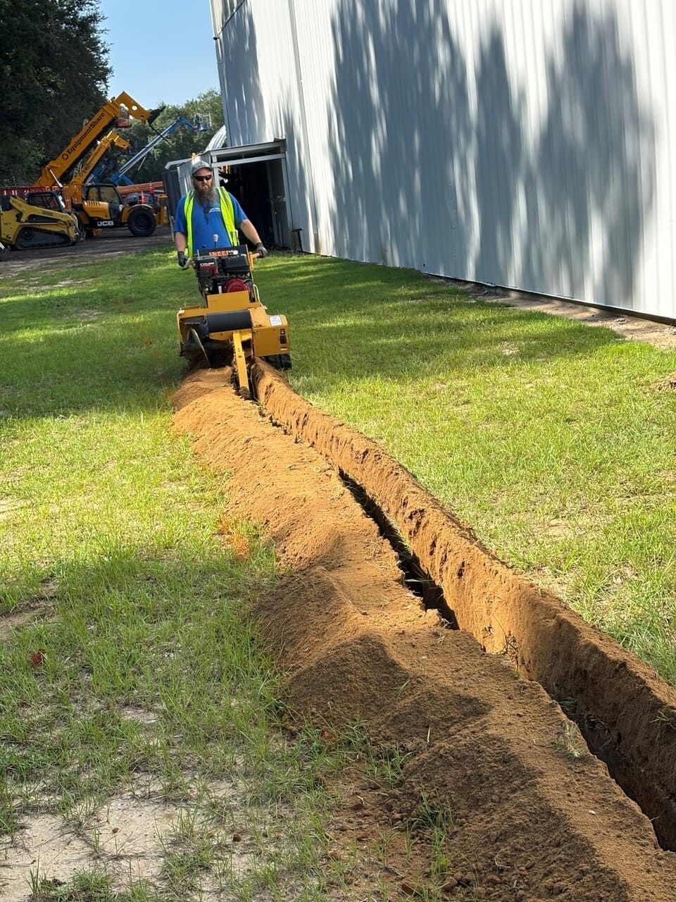 A worker operates a yellow trencher to dig a narrow channel through a grassy lawn beside a metal building.