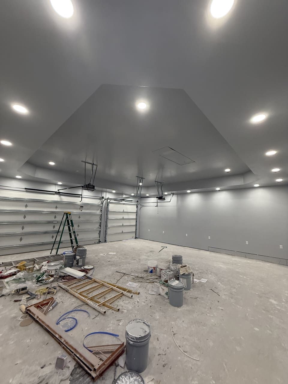 A spacious, unfinished gray room with recessed ceiling lights, an open garage door, a ladder, and construction buckets.
