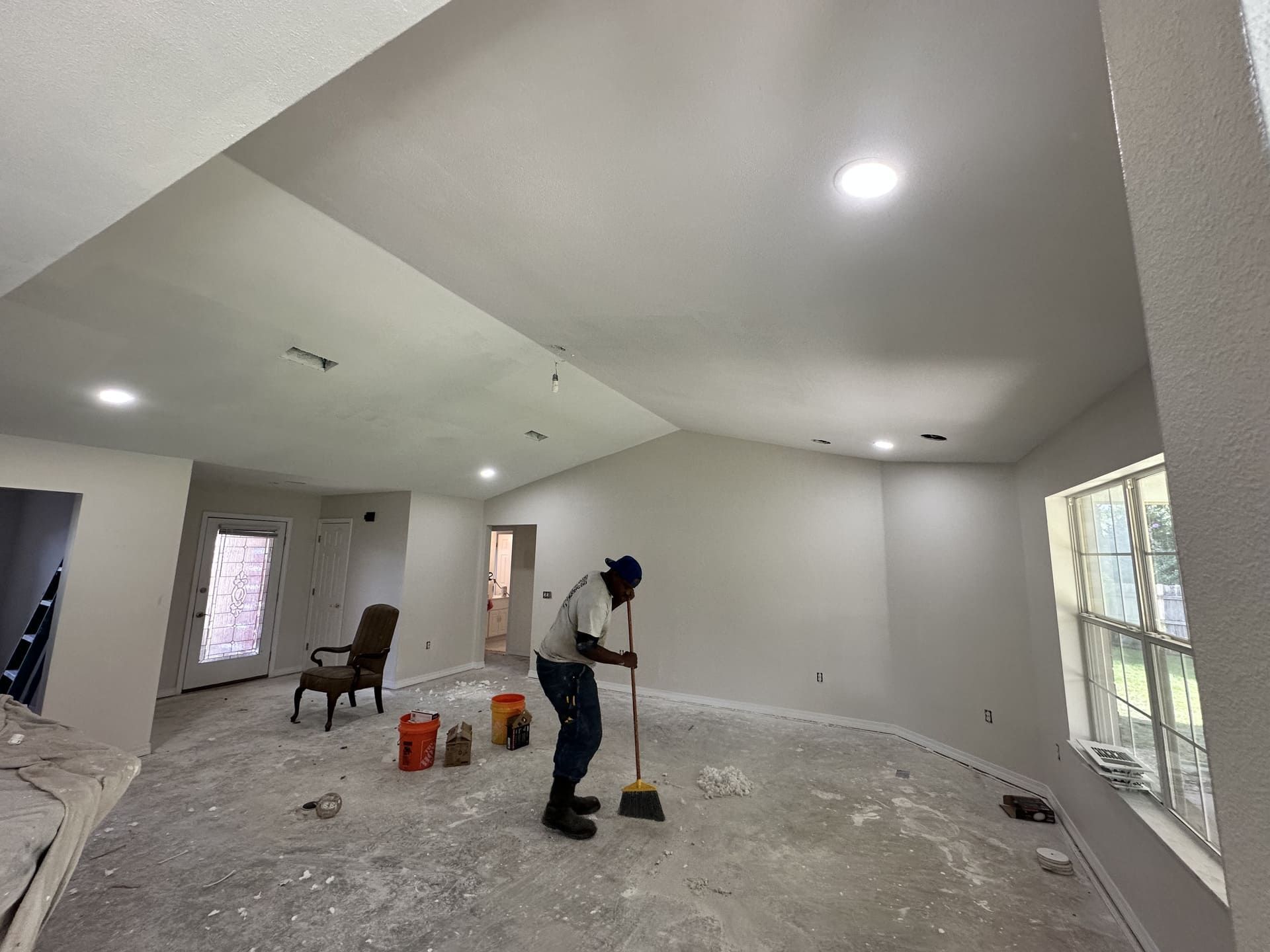 A person sweeps construction dust from the floor of a freshly painted, light-filled room with a vaulted ceiling.