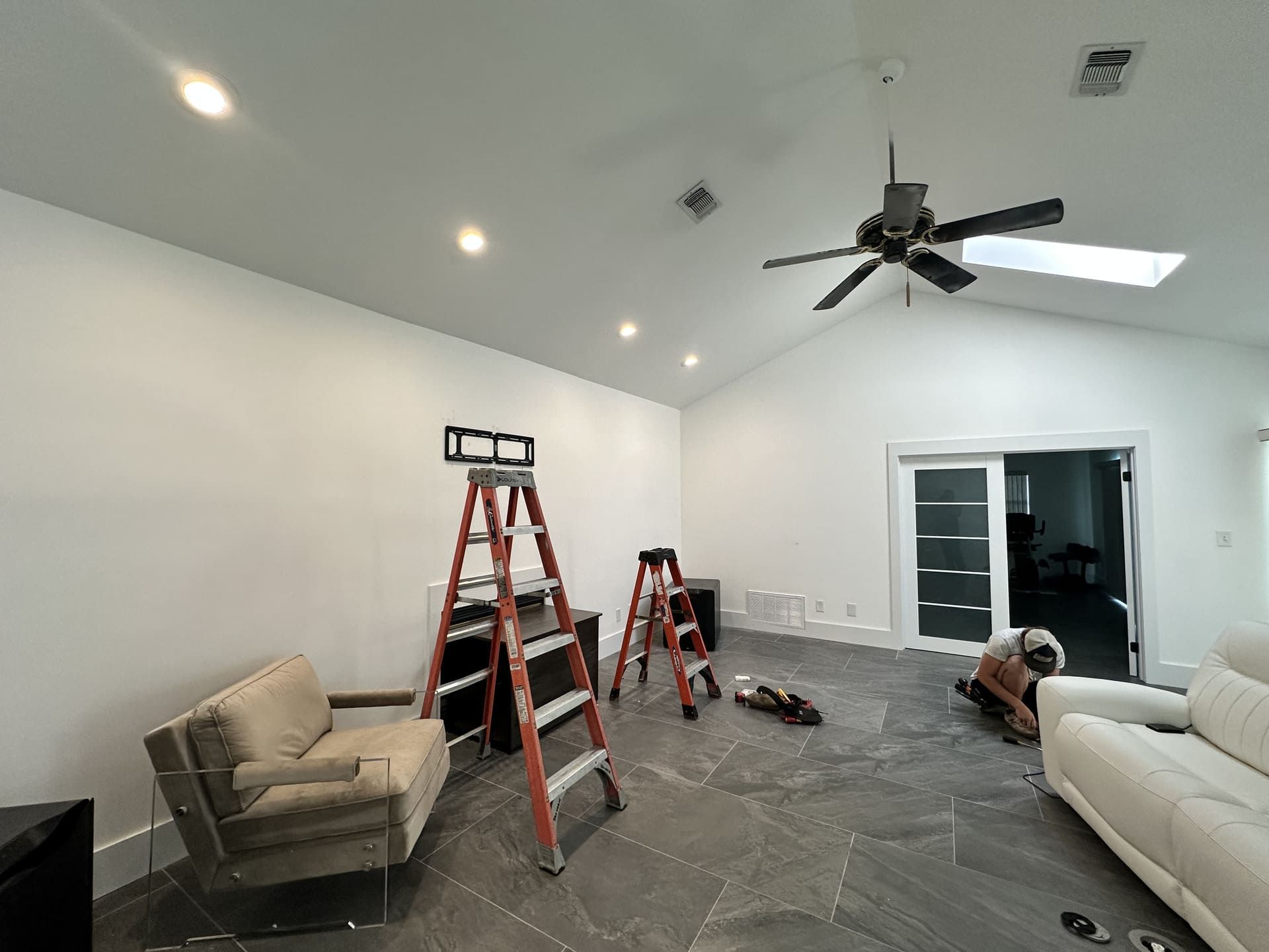 A room with a vaulted ceiling, gray tile floor, and white walls; a person works on the floor near two step ladders.