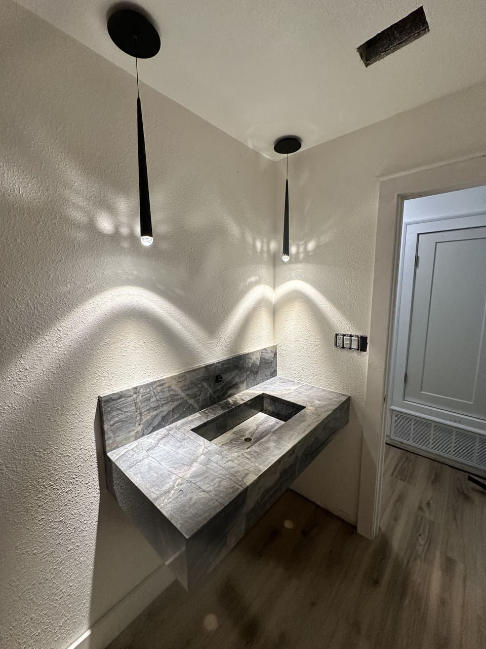 A floating grey stone bathroom sink mounted on a textured white wall, illuminated by two thin, black pendant lights.