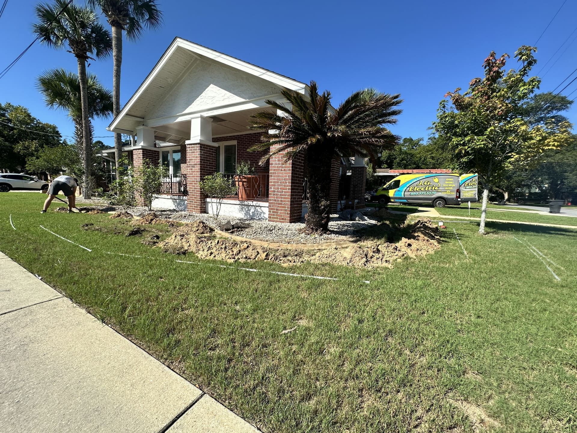 A person works in a residential front yard with dug-up dirt beds in front of a brick house with a white covered porch.