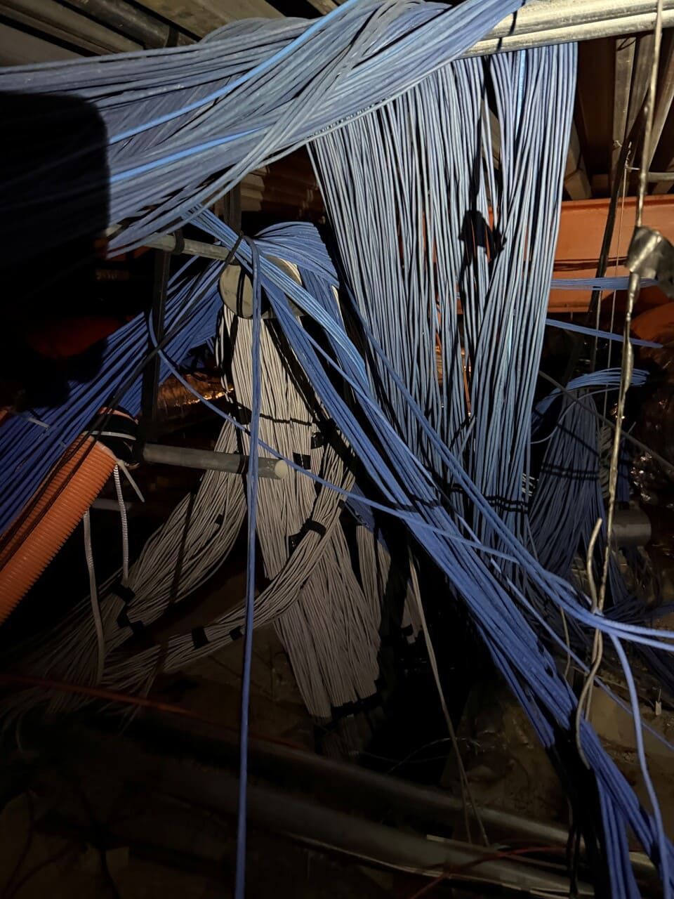 A dark, cluttered crawlspace featuring bundles of blue ethernet cables hanging from the ceiling near structural beams.