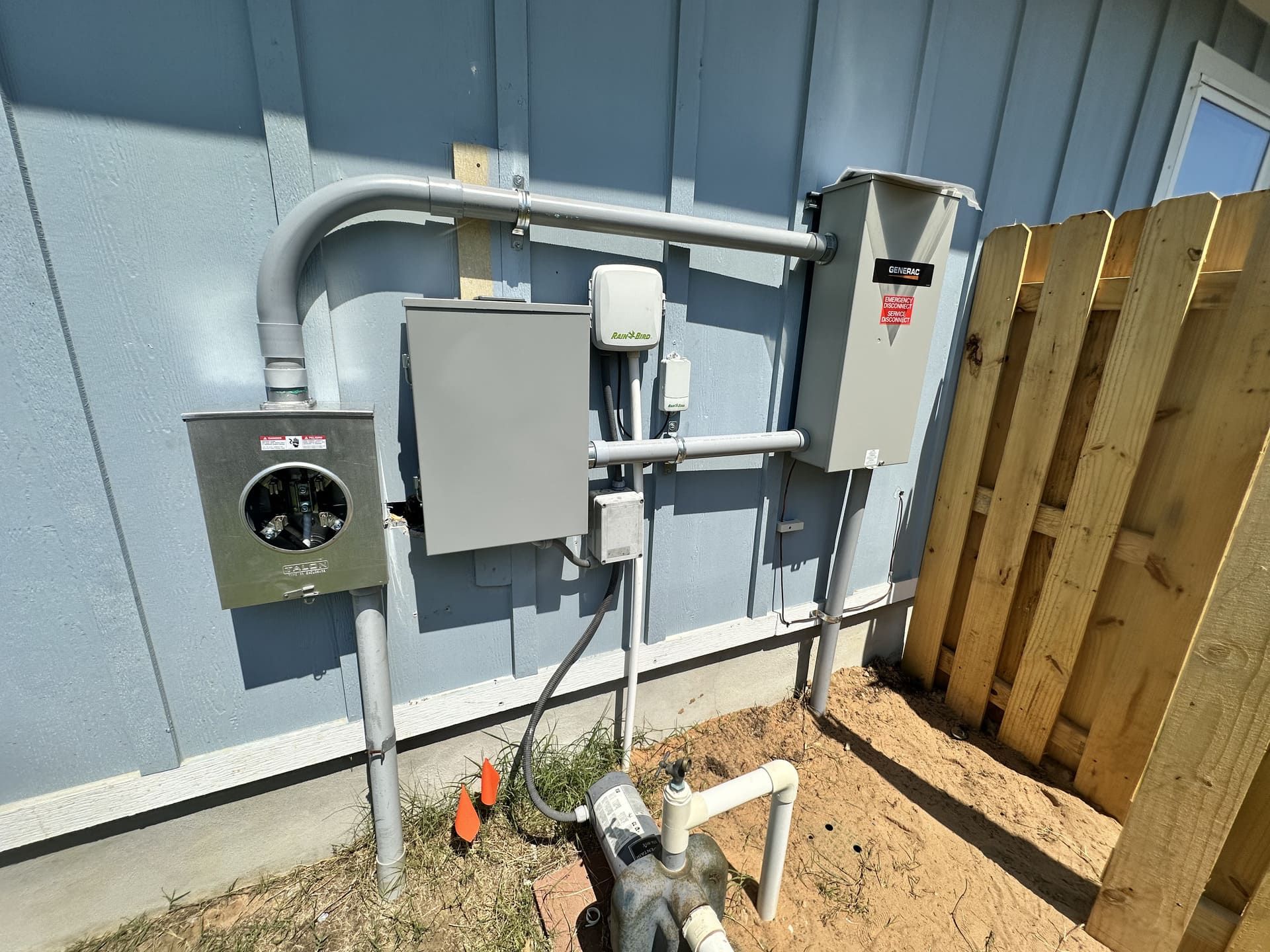 Exterior wall of a house with an electric meter, utility boxes, and pipes next to a wood privacy fence.
