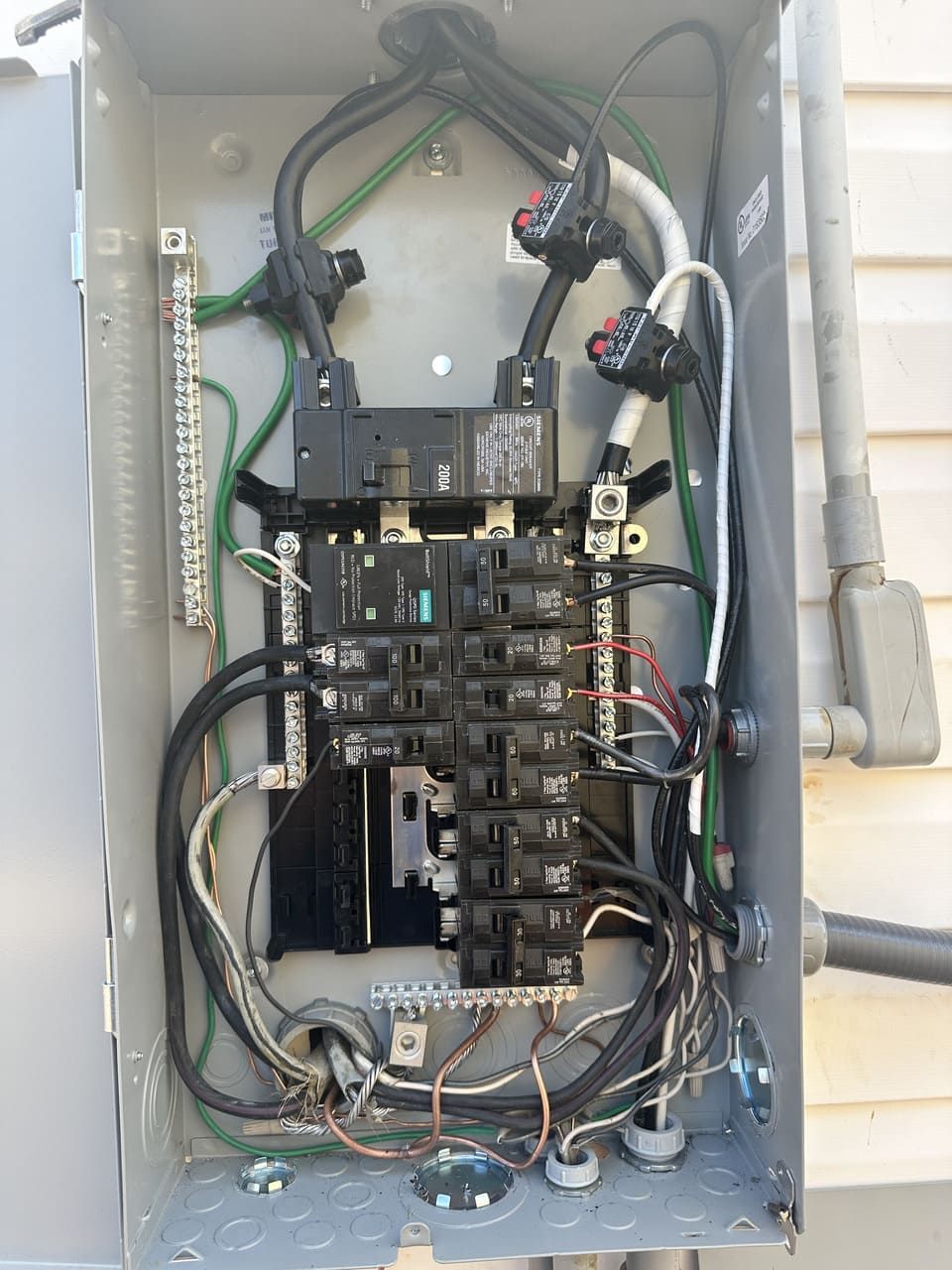 An open electrical service panel showing breakers, wiring, and metal buses housed in a gray metal enclosure on a wall.
