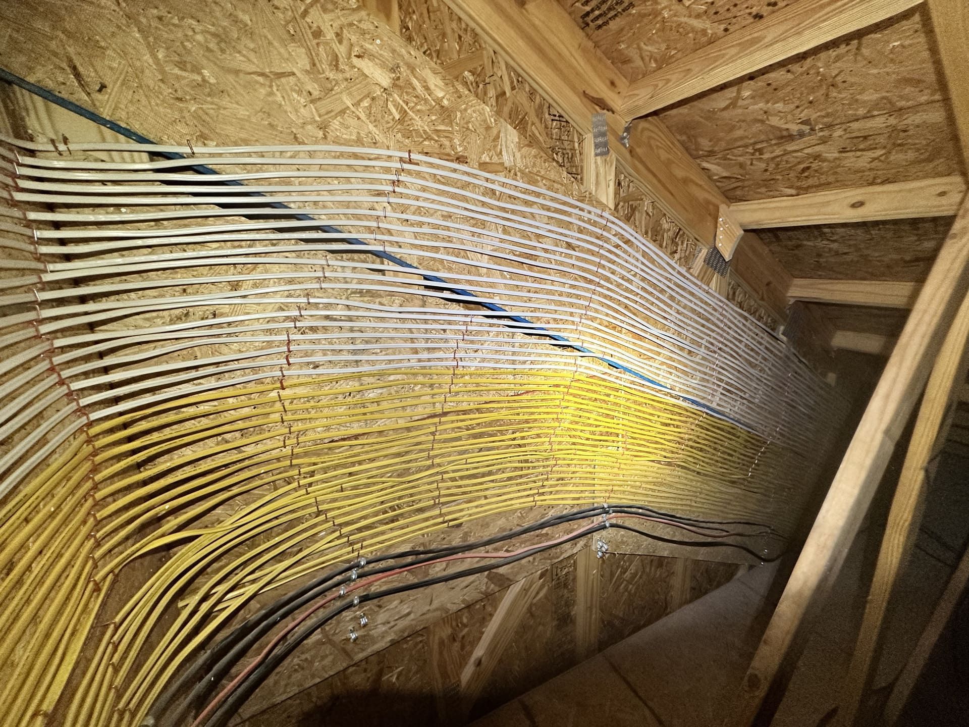 Numerous electrical cables in white, yellow, and black run in parallel along a wooden attic rafter.