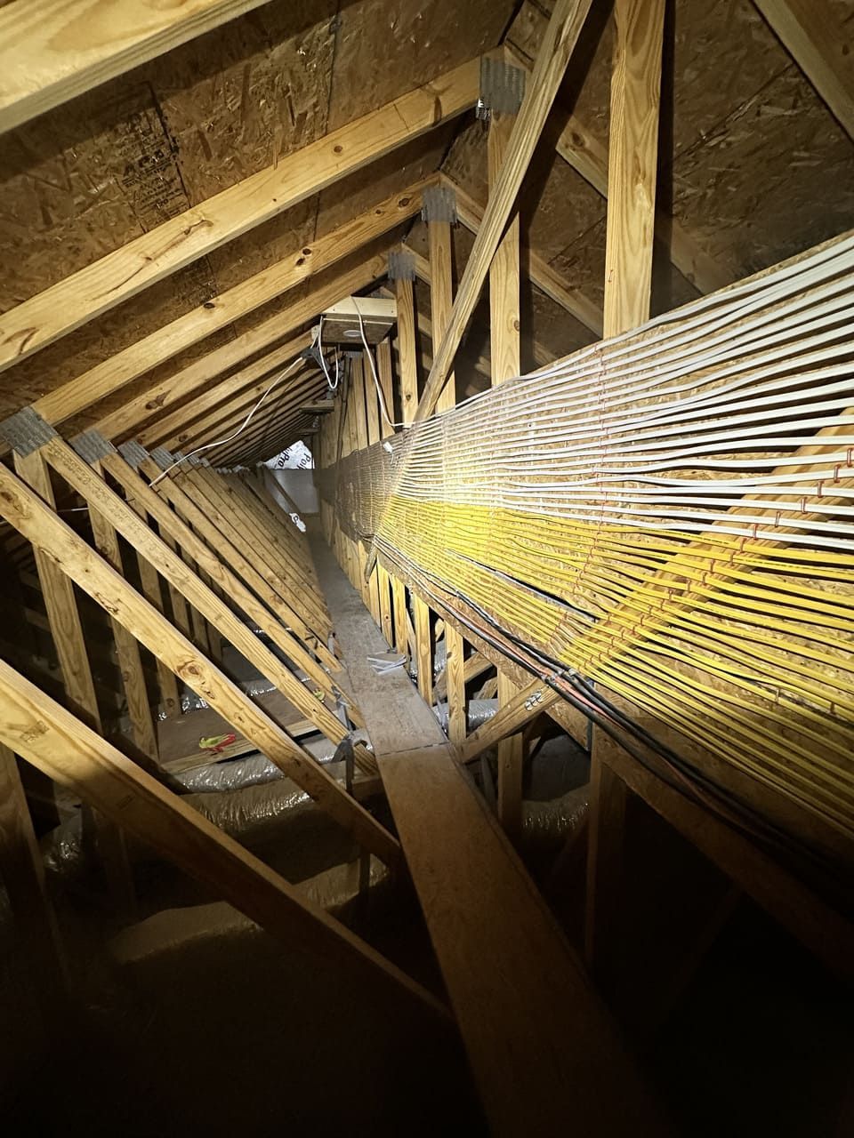 A view inside an attic, showing wooden roof trusses, a narrow wooden walkway, and a large, yellow-netted flexible duct.