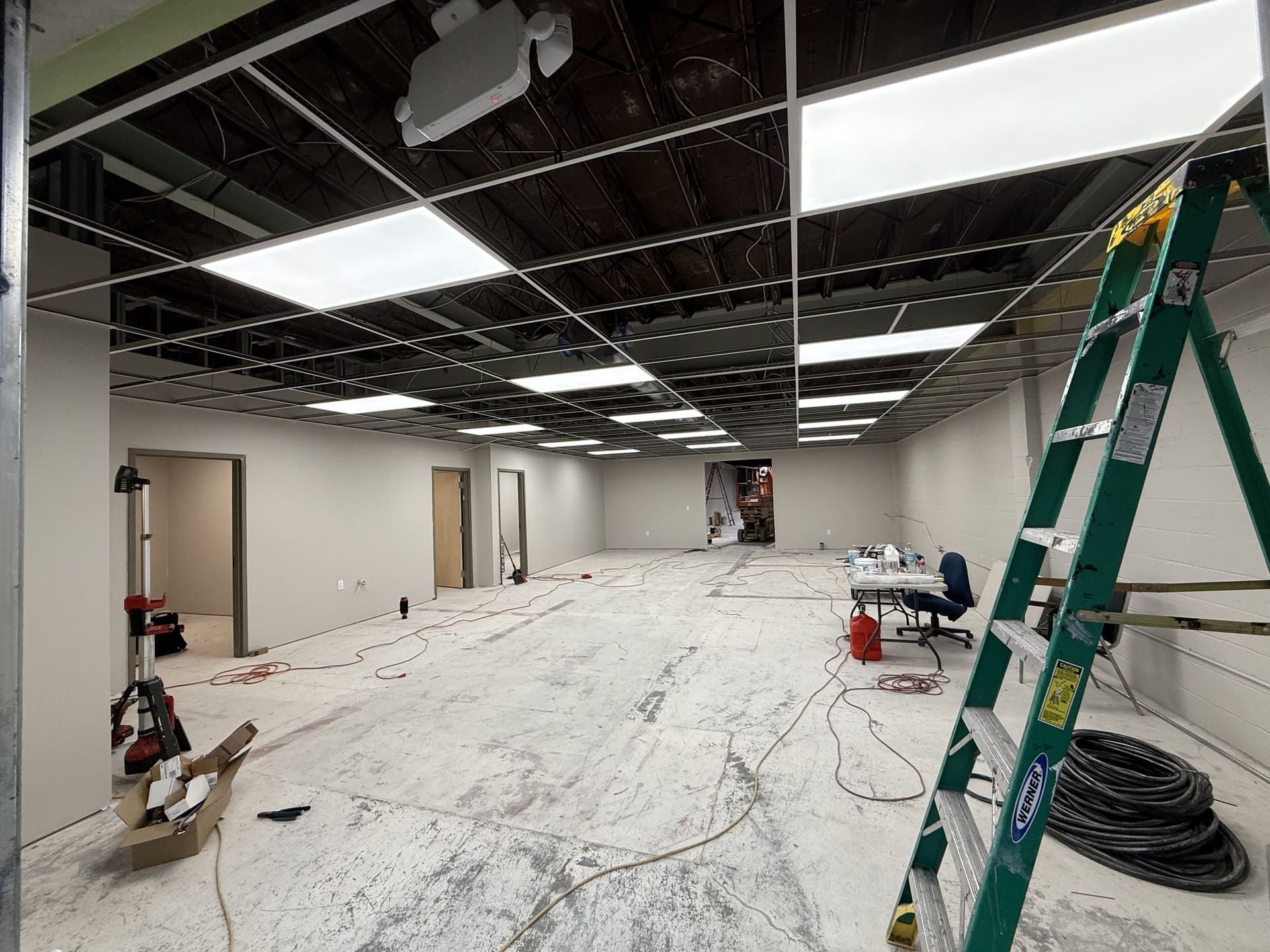 A commercial office space under construction with an exposed black ceiling, recessed lighting, and a green step ladder.