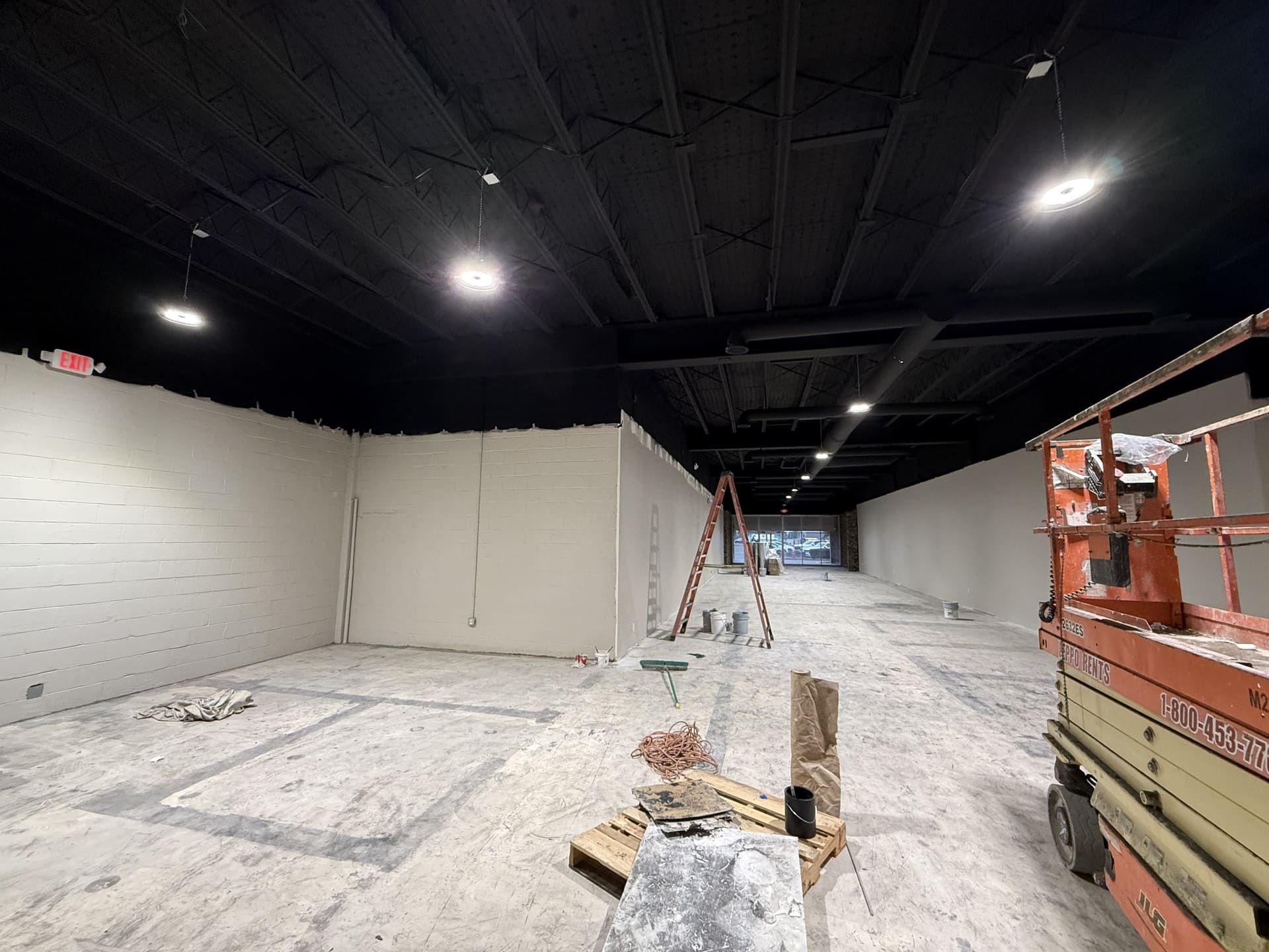 An indoor construction site with a high black ceiling, unfinished beige walls, concrete floors, and a scissor lift.