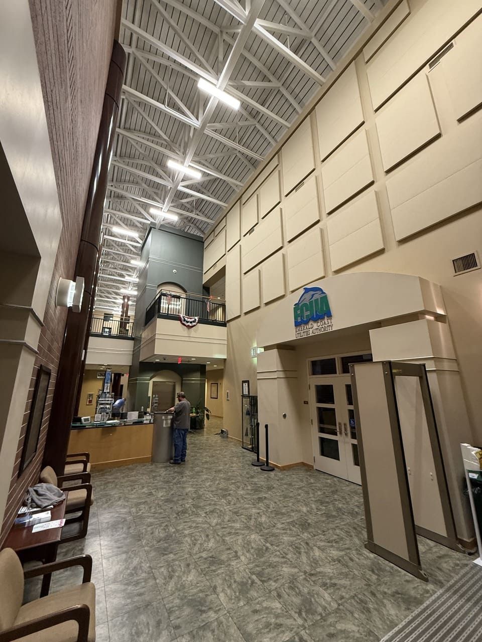 A spacious, high-ceilinged lobby with tan walls, tiled floors, a reception desk, and double doors under an arched sign.
